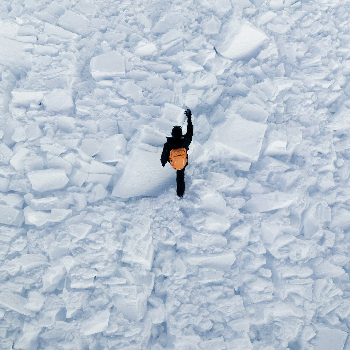 Blick von oben auf ein Lawinenfeld, ein Retter mit orangem Rucksack sucht zwischen den Schneeblöcken