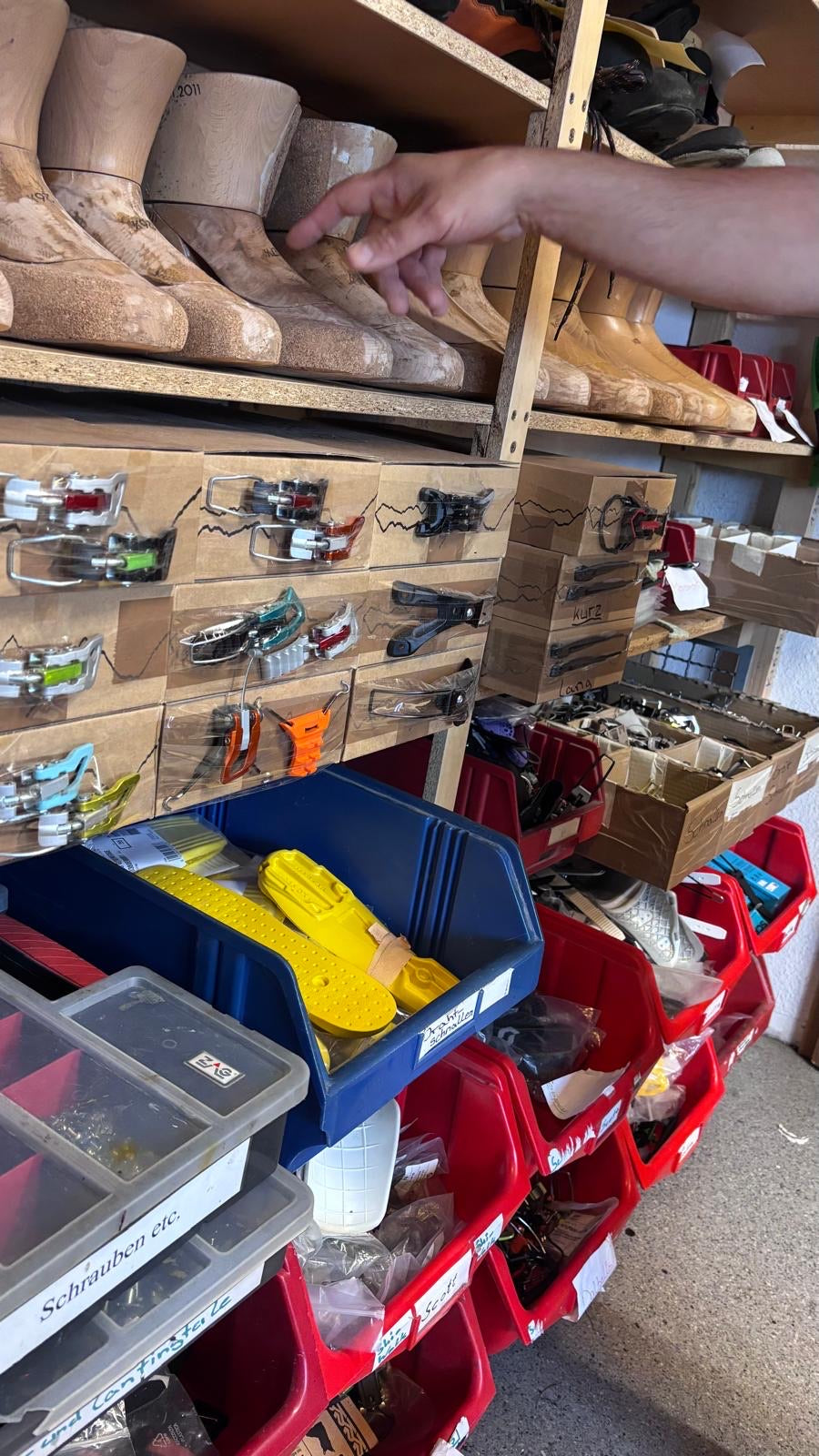 Regal in der Werkstatt mit Holzleisten in Reihen, darunter Schubladen voller Skischuhschnallen und Kisten mit Kleinteilen.