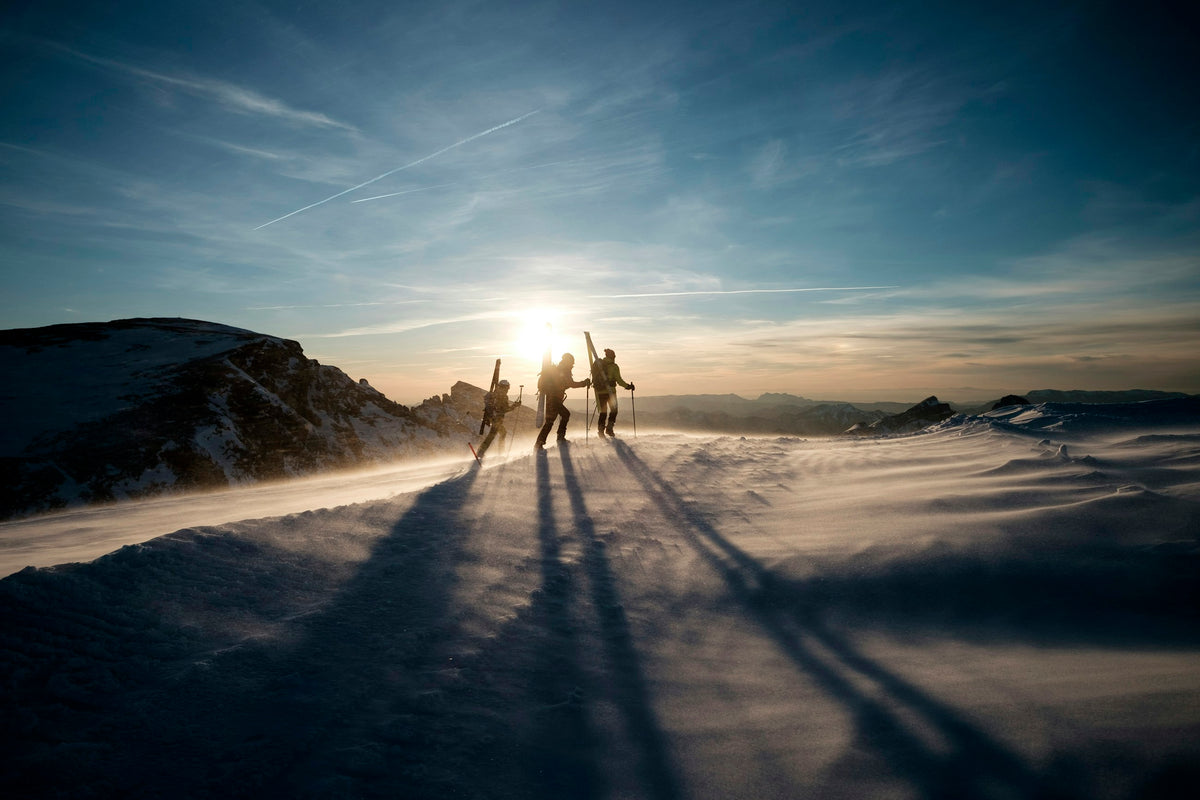 Drei Bergsportler tragen ihre Ski über einen verschneiten Bergrücken im Abendlicht der französischen Alpen.