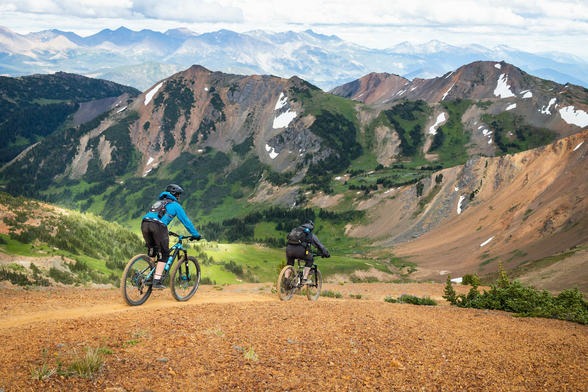 Zwei Mountainbiker auf einem schmalen Trail vor weitem Bergpanorama