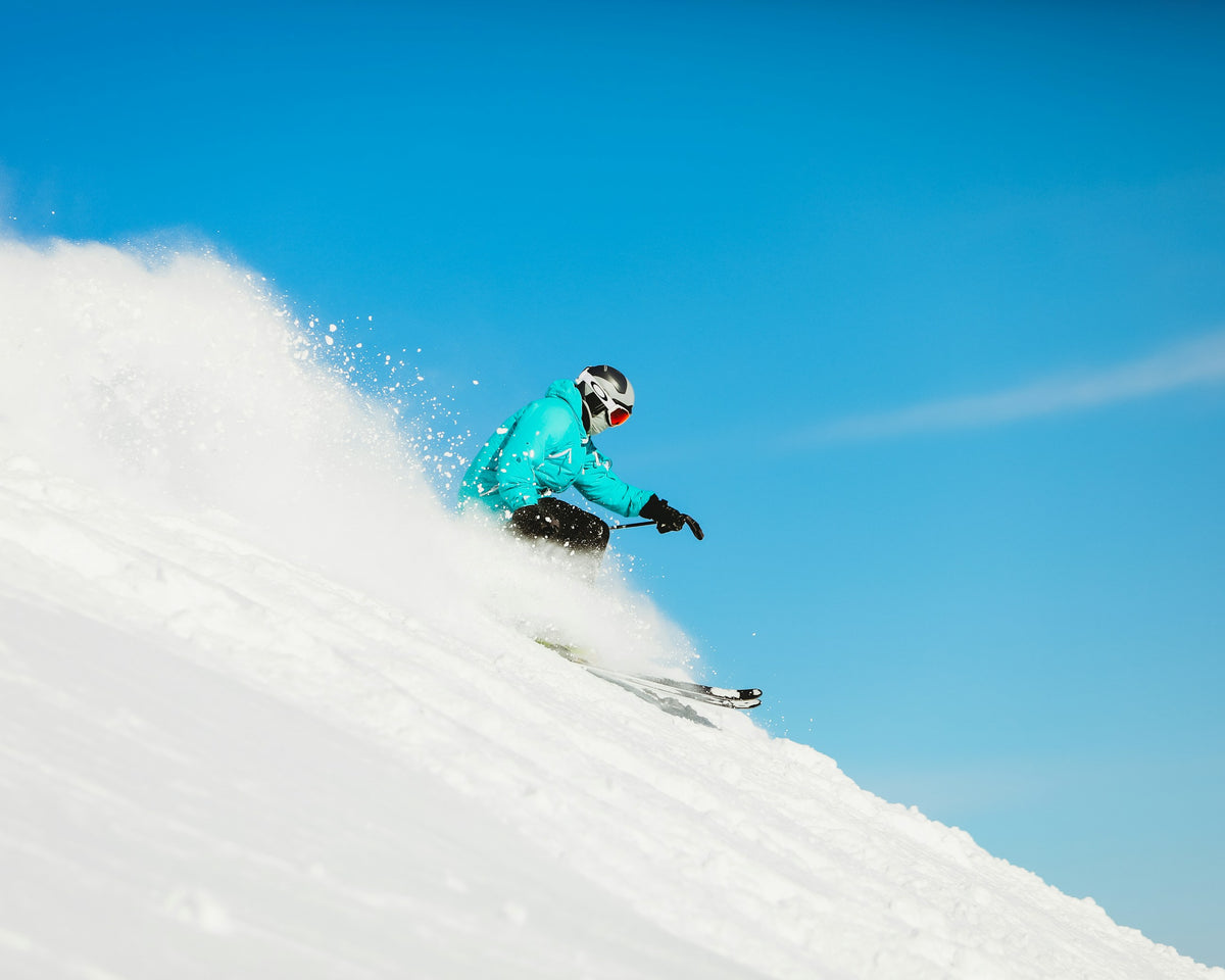 Freerider zieht mit Ski einen Schwung durch frischen Pulverschnee vor blauem Himmel.